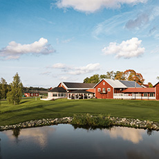 Vy över ett rött klubbhus på en golfbana med vattenhål med spegelblank yta i förgrunden.