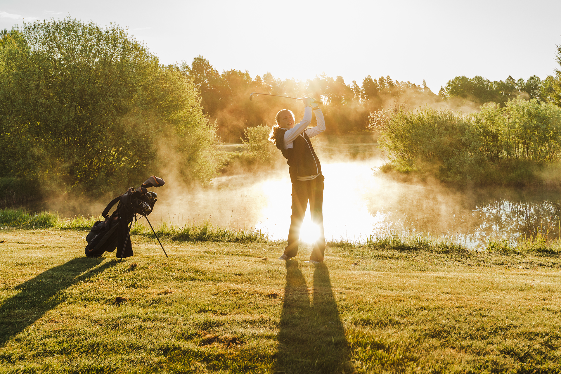 Tjej står och svingar i solnedgången på en golfbana, i bakgrunden syns en damm.