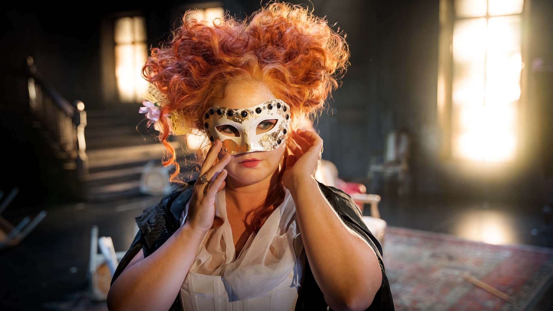 Skådespeleska med rött yvigt hår håller en teatermask för ansiktet. Foto Markus Gårder.