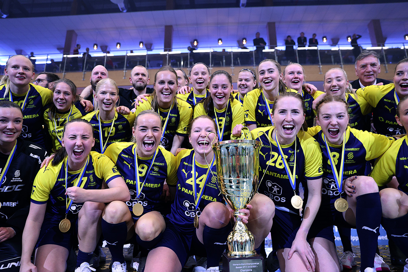 Gruppbild på det svenska damlandslaget i innebandy. De håller fram en guldpokal mot kameran och ler.