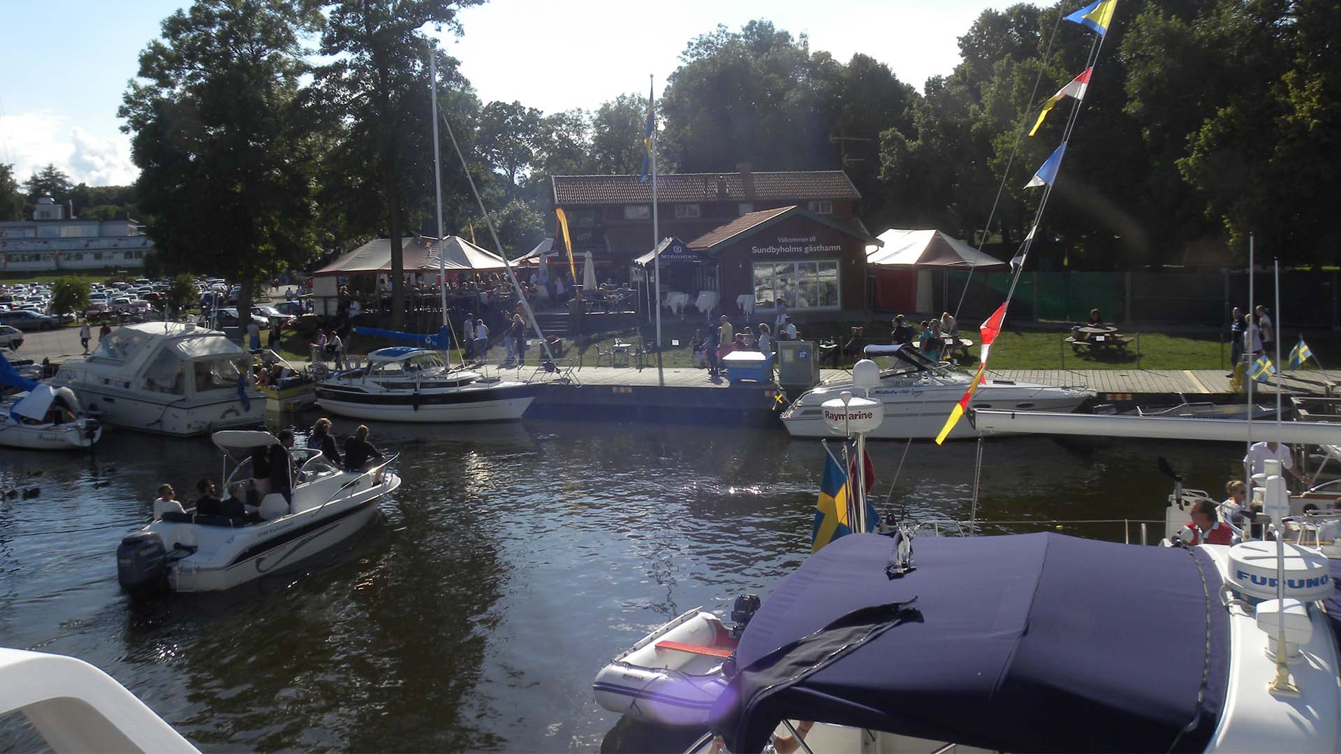 Båtar i en gästhamn med restaurang full med folk på uteserveringen. Båtar i en gästhamn med restaurang full med folk på uteserveringen.