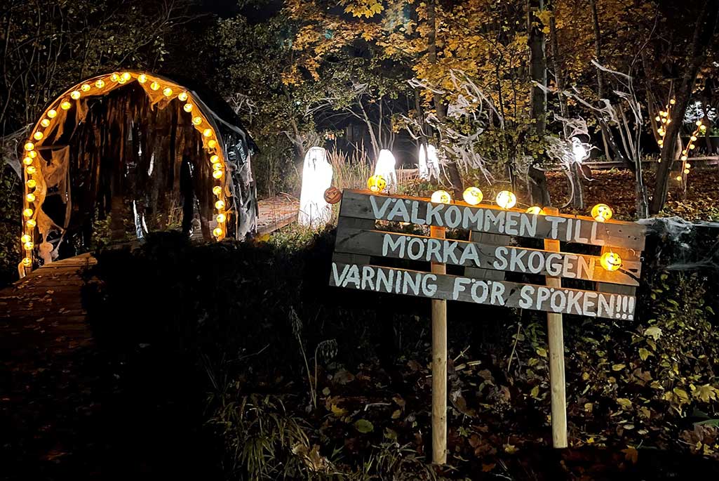 En skylt med texten "Välkommen till mörka skogen! Varning för spöken" med en mörk skog i bakgrunden En skylt med texten "Välkommen till mörka skogen! Varning för spöken" med en mörk skog i bakgrunden