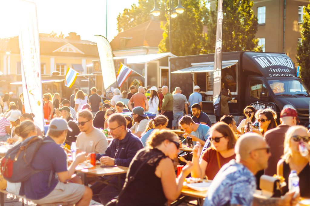 Människor sitter vid långbord och äter under evenemanget Västerås street food. Foodtrucks står i bakgrunden.