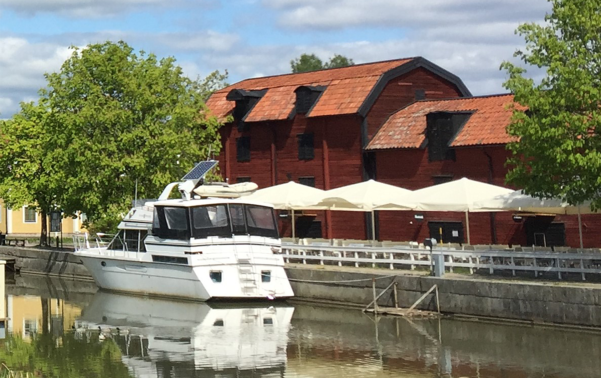 Båt ligger vid kaj i Enköpings gästhamn. I bakgrunder syns uteservering framför magasinbyggnader. Pressbild