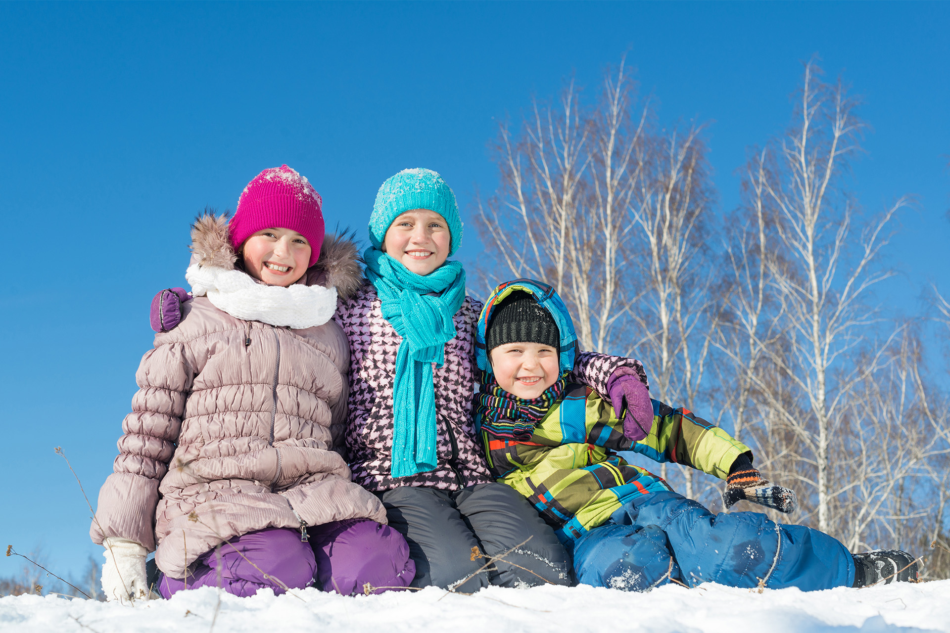 Tre glada barn sitter i snön en strålande dag. Foto: Mostphotos
