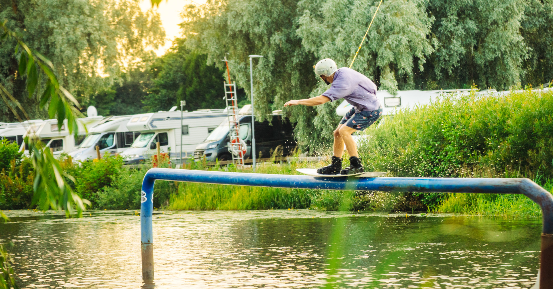 Wakeboardåkare glider över en rail i vattnet vid Lögarängen i Västerås, med husbilar och grönska i bakgrunden. Wakeboardåkare glider över en rail i vattnet vid Lögarängen i Västerås, med husbilar och grönska i bakgrunden.
