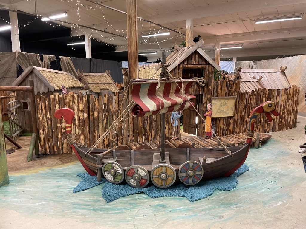 Vy över interiör till en barnutställning om vikingaliv, i förgrunden en vikingabåt och små byggnader i bakgrunden.