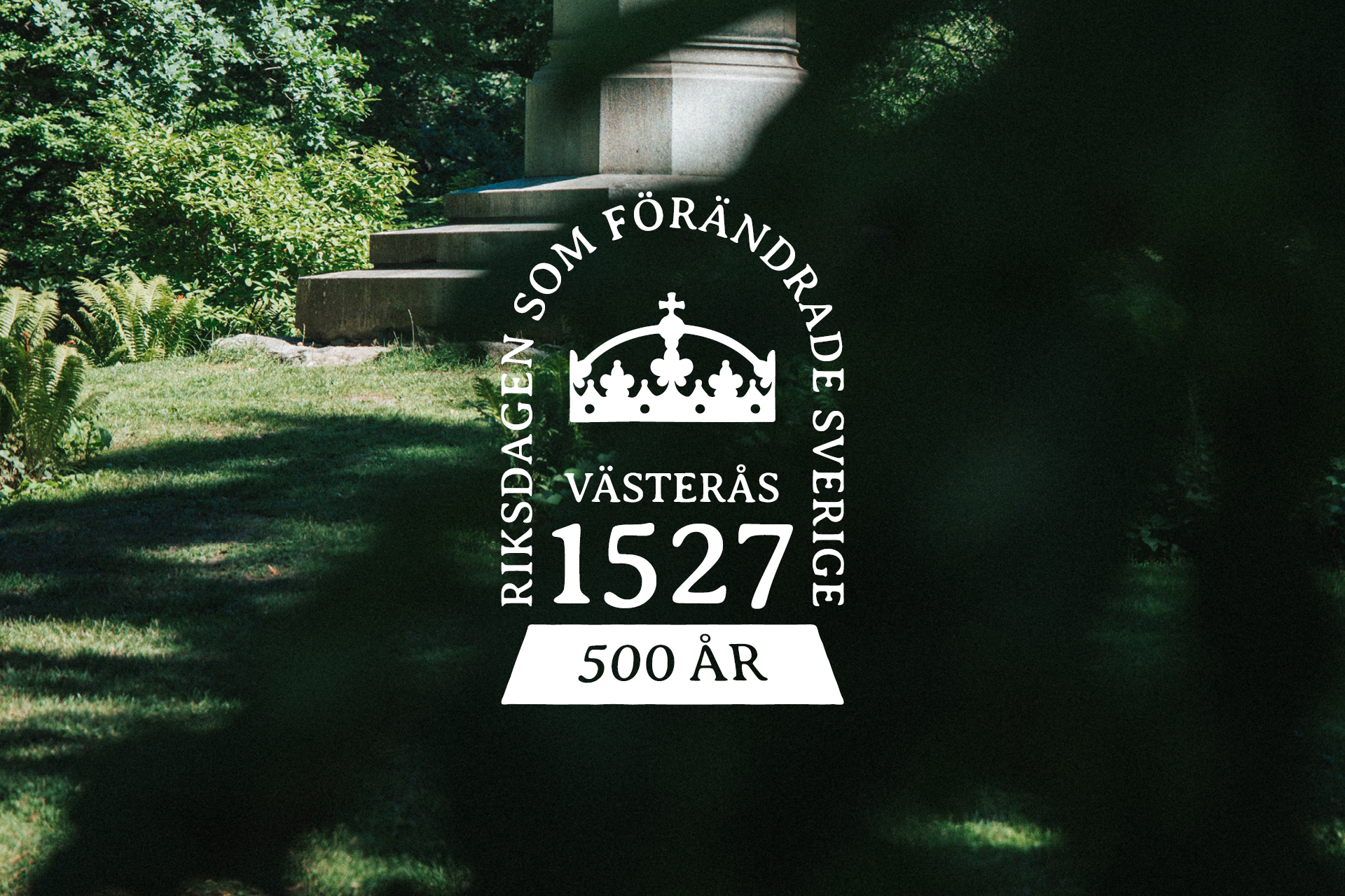 Västerås 1527-logotyp mot grönskande bakgrund i Vasaparken.