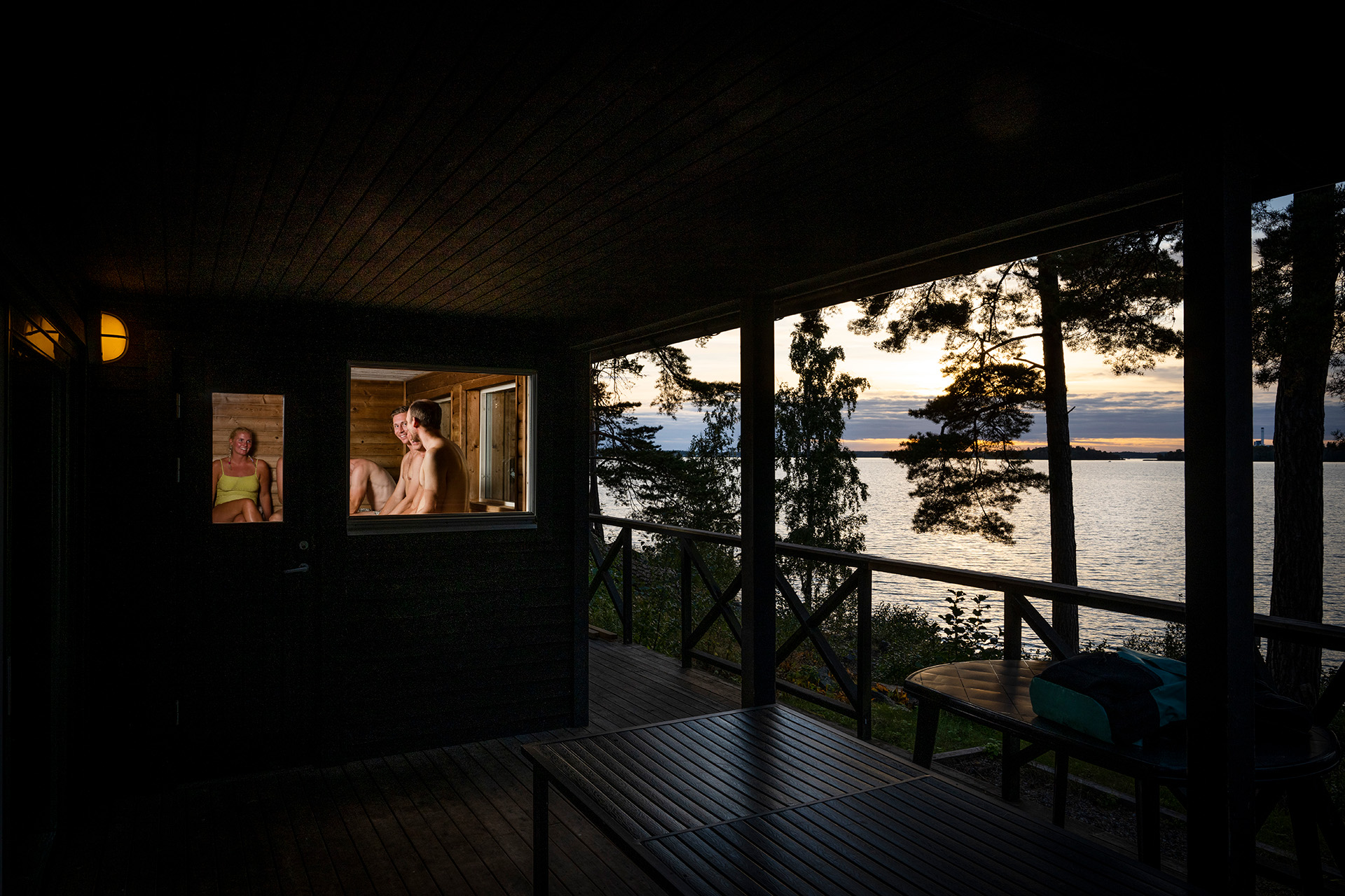 Genom ett fönster ser man tre kompisar i en bastu med sjöutsikt.