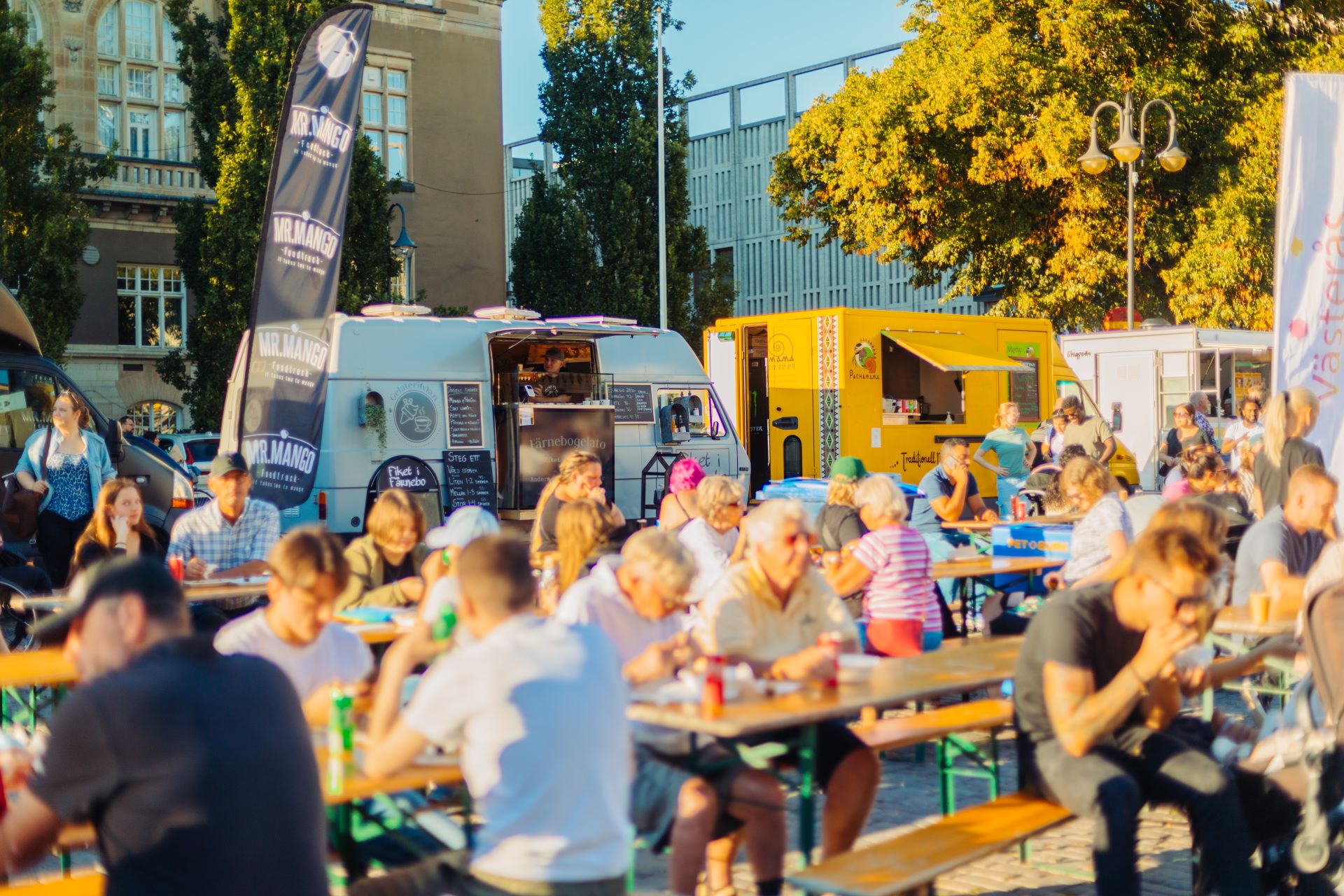 Folksamling på ett soligt torg där människor sitter vid långbord och äter, med flera foodtrucks i bakgrunden