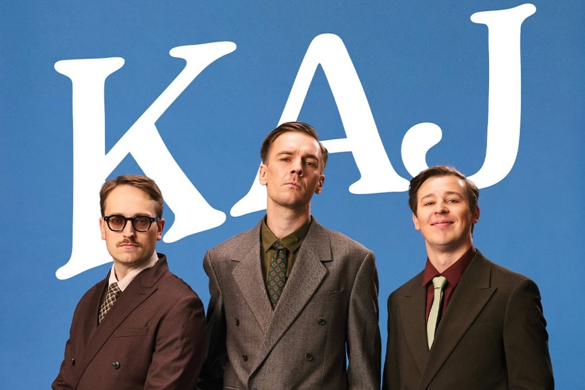 Medlemmarna i KAJ står framför en blå bakgrund, i bruna kostymer under en text som lyder "KAJ".