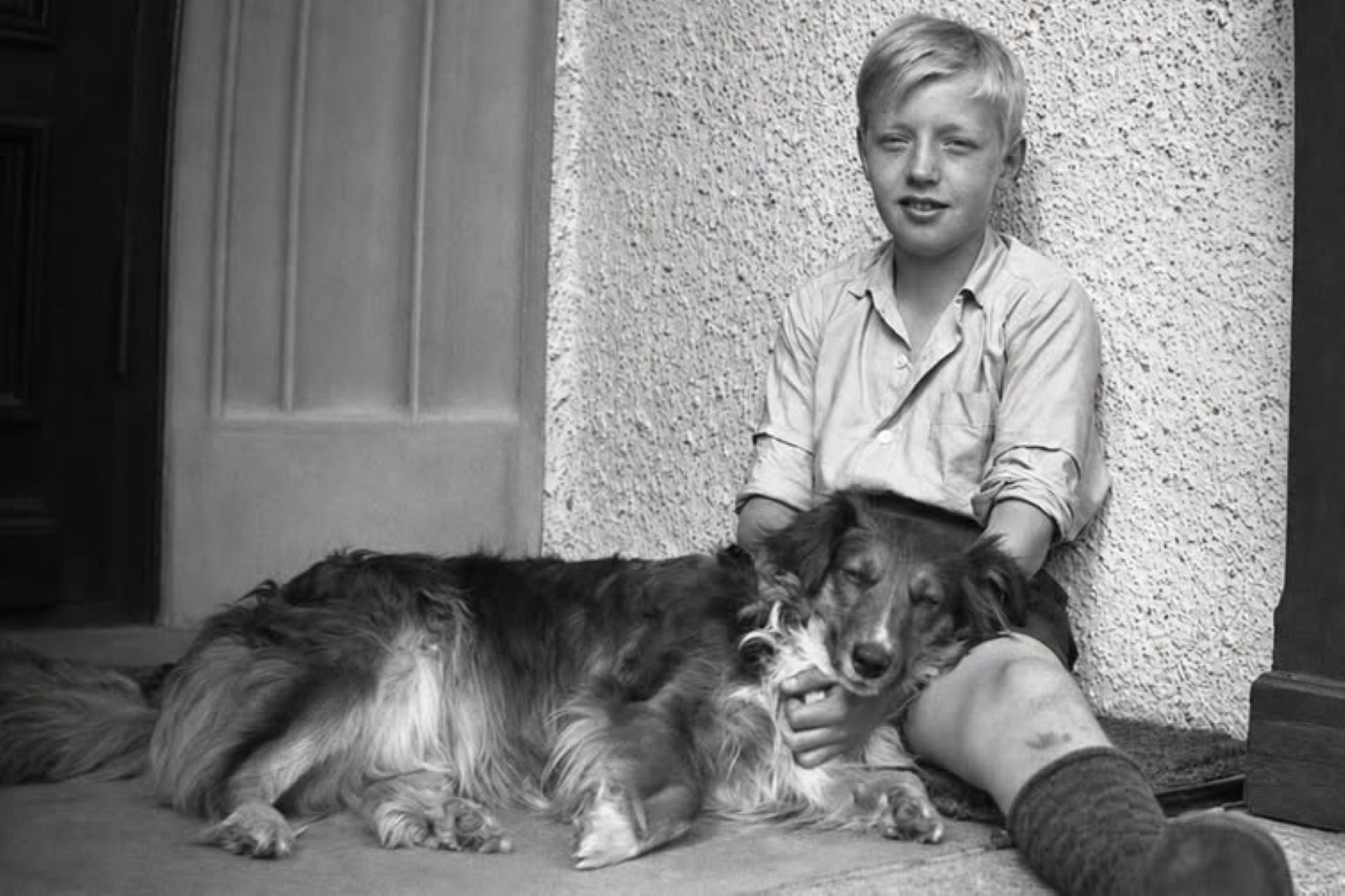 Ett svartvitt fotografi föreställande en pojke lutad mot en vägg med en hund liggandes i hans knä.