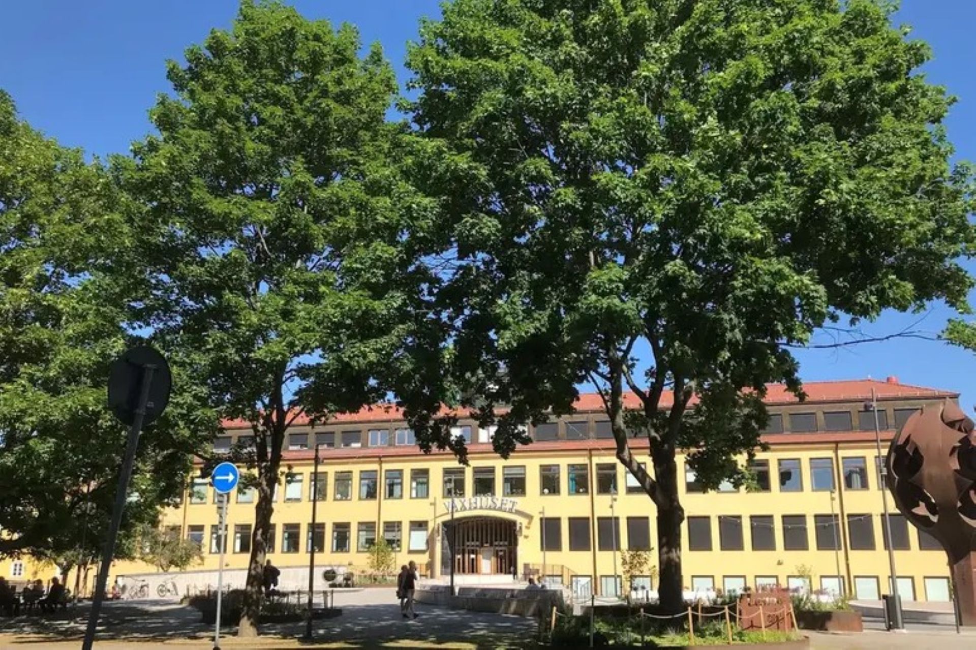 Exteriörbild på ett soligt Växhuset med gröna träd i förgrunden.